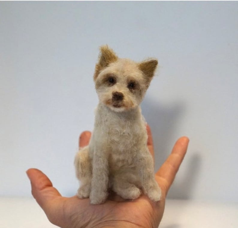 May include: A small, white, needle felted dog with brown eyes and a light brown muzzle. The dog is sitting on a person's hand.