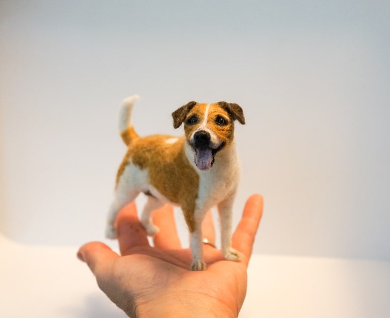 May include: A needle felted dog figurine, a Jack Russell Terrier, with brown and white fur, standing on a hand. The dog has a brown nose, black eyes, and a pink tongue sticking out.