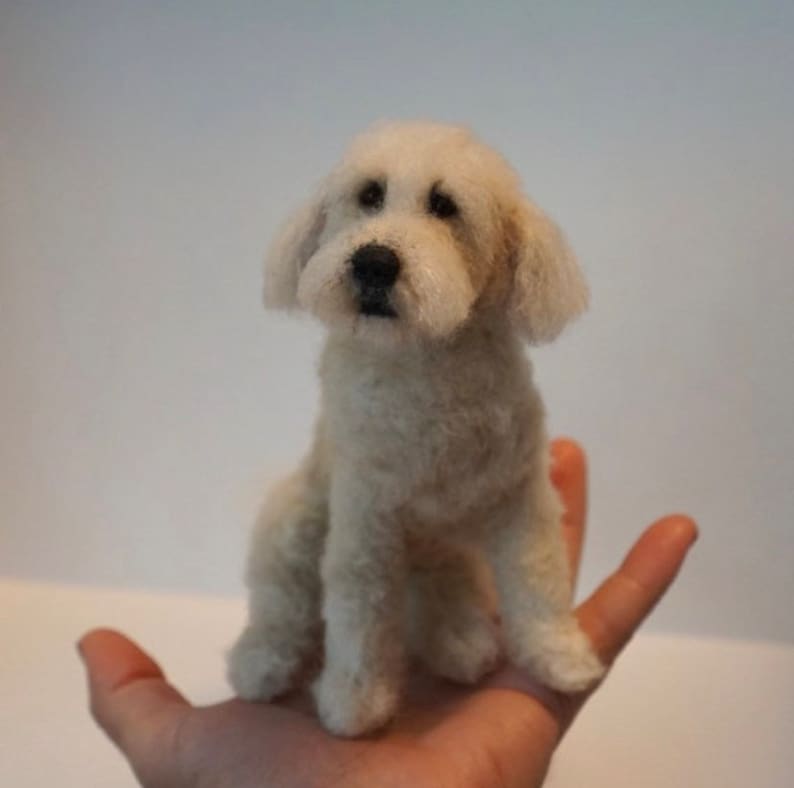May include: A needle felted dog sculpture, a white and cream colored miniature dog with black eyes and a black nose, sitting on a hand.