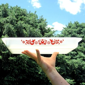 May include: A rectangular white glass baking dish featuring a vibrant red pattern of stylized birds and floral motifs centered along the side. The dish has integrated handles on both ends and a smooth, semi-translucent finish throughout the body.