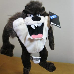 May include: A plush Taz, the Tasmanian Devil, bean bag toy. The cartoon character has a white belly, black mouth, and a pink tongue. The toy has a black tag that says "BEAN BAG".