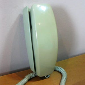 May include: A vintage, wall-mounted telephone in a pale green color. The phone has a rectangular shape with a curved front and a coiled handset cord. The phone is mounted on a light-colored wooden surface.