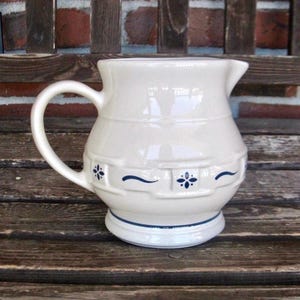 May include: A cream-colored ceramic pitcher featuring a rounded body with a handle and a pouring spout. The exterior is decorated with a repeating blue pattern of small four-petaled floral motifs and horizontal wavy lines around the center.