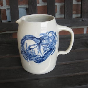 May include: Cream-colored ceramic pitcher with a blue abstract design. The pitcher has a handle and a spout for pouring. The blue design appears to be hand-painted and is centered on the front of the pitcher.