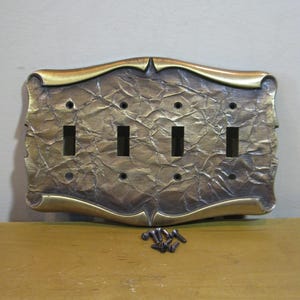 May include: A rectangular four-gang light switch wall plate with a metallic bronze finish and a textured, crinkled surface. The plate features smooth, curved edges and includes eight small dark metal screws for installation purposes.