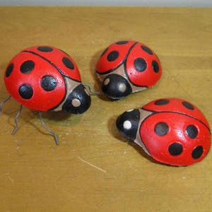 May include: Three decorative ladybug figurines. Each ladybug has a red body with black spots, a black head, and a tan-colored section. One ladybug has wire legs.