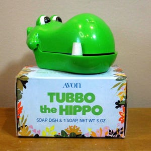 May include: A vintage green plastic Tubbo the Hippo soap dish by Avon, with a white soap piece inside. The box below is light blue with floral designs and the text "TUBBO the HIPPO".
