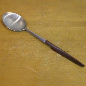 May include: A stainless steel serving spoon with a dark brown wooden handle. The spoon has a large, oval bowl and a long, slender handle. The handle is made of dark brown wood and is attached to the spoon with a silver metal piece.