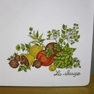 May include: A white rectangular surface featuring a colorful botanical illustration of various vegetables, nuts, and herbs, including a mushroom, tomato, and artichoke. The text "La Sauge" is printed in a cursive black font in the bottom right corner.