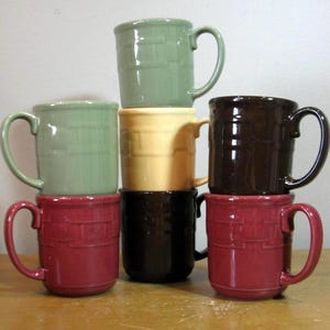 May include: A stack of six ceramic mugs in various colors including sage green, cream, dark brown, and deep red. Each mug features a consistent embossed basket-weave pattern around the exterior, a smooth glossy finish, and a curved side handle.