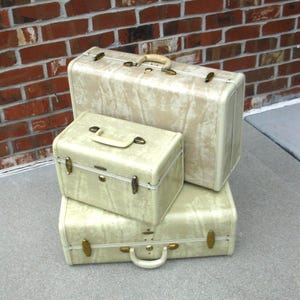 May include: A set of three off-white suitcases with a marbled texture and brass-toned hardware. The collection includes one large rectangular suitcase, one medium rectangular suitcase, and one smaller train case, all featuring top handles and latches.