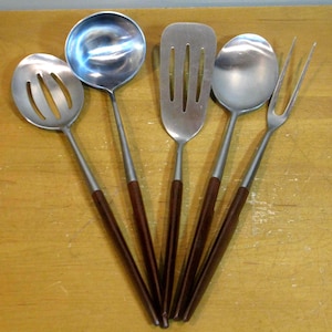 May include: A set of five stainless steel kitchen utensils with dark brown wooden handles. The set includes a slotted spoon, ladle, spatula, solid spoon, and serving fork. The utensils are arranged on a light brown wooden surface.