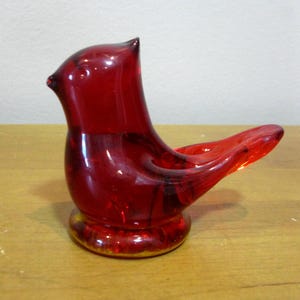 May include: A vibrant red glass bird figurine with a rounded base and a curved tail. The bird has a detailed beak and a small, pointed crest. The figurine is made of transparent red glass.