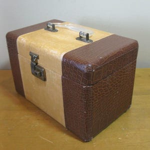 May include: A vintage rectangular travel case with a light tan center and dark brown faux-crocodile skin trim. It has a clear plastic handle and a metal clasp closure. The case is likely from the mid-20th century.