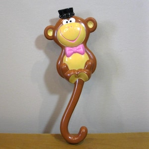 May include: A brown monkey-shaped umbrella handle with a yellow face, pink bow tie, and black top hat. The handle has a curved, hook-like design. The monkey's face has a smiling expression.