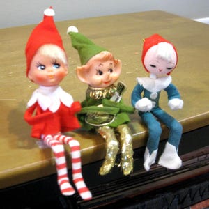 May include: Three elf figurines with painted faces. The first wears a red hat and red outfit with white striped legs. The second wears a green hat, green outfit, and gold pants. The third wears a red hat and a blue outfit with white trim.
