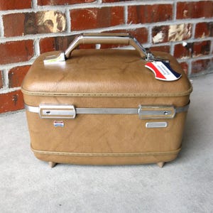 May include: A tan hardshell train case with a textured finish, featuring a silver-tone metal handle, two locking latches, and a small tag attached to the handle that reads American Tourister in red, white, and blue colors on a white background.