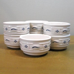 May include: A set of six white ceramic bowls. Each bowl features a repeating pattern of a blue curved line and a four-petal flower. The bowls are stacked and arranged on a light brown surface.