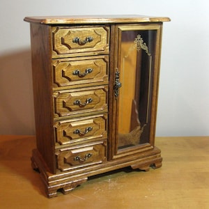 May include: A wooden jewelry armoire with five drawers on the left and a glass-door cabinet on the right. The armoire is made of light brown wood with ornate drawer pulls and decorative glass etching.