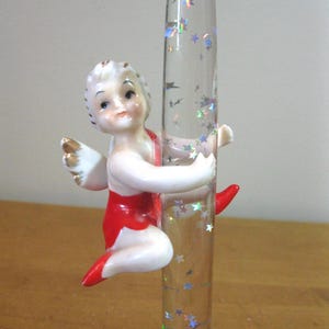 May include: A porcelain cherub figurine with a red and gold costume is climbing a clear glass tube filled with iridescent glitter stars.
