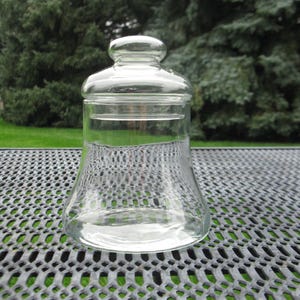 May include: A clear glass jar with a bell-shaped body and a matching removable glass lid. The lid features a rounded knob handle at the top. The jar has a wide base that tapers slightly toward the neck, providing a smooth, transparent surface.