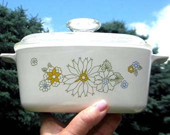 Op de afbeelding: Een rechthoekige witte ovenschaal met een doorzichtig glazen deksel en geïntegreerde handgrepen aan de zijkanten. De buitenkant is voorzien van een bloemmotief in geel, blauw en groen, met gestileerde madeliefjes en bladeren in het midden.
