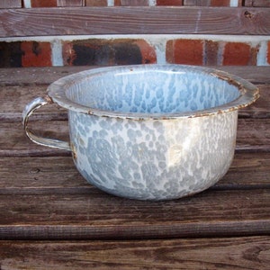 May include: A vintage white and blue speckled enamelware bowl with a single handle. The bowl has a rustic, worn look and is perfect for farmhouse decor.