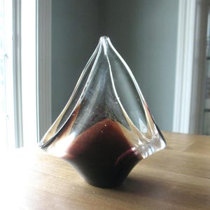 May include: A clear glass decorative bowl with a unique shape. The bowl has a dark burgundy base that fades to a clear top. The bowl is sitting on a wooden surface.