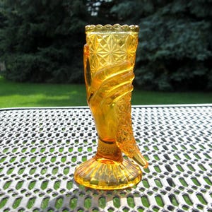 May include: An amber-colored glass vase shaped like a hand holding a cornucopia. The vessel features a textured diamond pattern on the upper section and a scalloped rim. The base is circular with a ribbed design, supporting the hand-shaped structure.