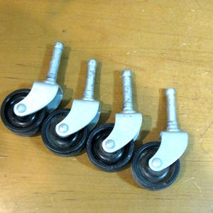 May include: Four caster wheels with black wheels, silver stems, and light gray brackets. The wheels are arranged in a row, angled slightly towards the right. The background is a light brown wooden surface.