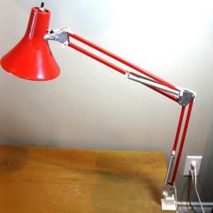 May include: A red desk lamp with a conical shade and adjustable arm. The lamp has a silver-colored joint and a white clamp to attach to a desk. The lamp is on a wooden surface.