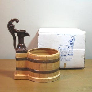 May include: A ceramic planter shaped like a wooden barrel with a dark brown water pump attached to the side. The barrel has light tan wood grain detailing and dark brown bands. A white cardboard box with a blue illustration is positioned behind it.