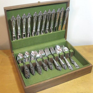 May include: A wooden storage chest with a green felt interior containing a set of silver-toned flatware. The collection includes various spoons, forks, and knives, each featuring an ornate, embossed handle design arranged in dedicated slots.