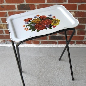 May include: A rectangular white metal tray table with a floral bouquet print featuring red, yellow, and purple flowers with green leaves. The tray has a scalloped edge and is supported by a black metal folding stand with four legs.