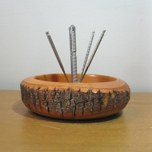 May include: A wooden nut bowl with a bark-like exterior and a smooth interior. It holds a silver nutcracker and three silver picks. The bowl sits on a light brown surface.