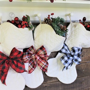 May include: Three white quilted Christmas stockings with different colored bows. The stocking on the left has a red and black plaid bow, the middle stocking has a red, black, and tan plaid bow, and the stocking on the right has a black and white checkered bow.