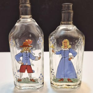 May include: Two clear glass bottles with silver-toned metal caps, each featuring a hand-painted figure. The left bottle depicts a person in a blue jacket, red shorts, and a brown hat holding a glass. The right bottle shows a person in a blue dress.