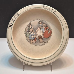 May include: Cream-colored ceramic baby plate with a green rim and a decorative illustration of children around a table. The plate has the words "BABY'S PLATE" and "Little Jack Horner, sat in a corner, eating his Christmas pie."