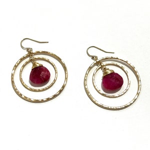May include: Gold hoop earrings with a single red gemstone dangling from each hoop.