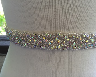 Op de afbeelding: Een decoratieve riem met iriserende kristallen en transparante kralen. De riem heeft een gevlochten ontwerp, waarbij de kristallen het licht vangen. Het accessoire wordt getoond tegen een witte achtergrond, wat suggereert dat het een modeartikel is.