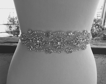 Op de afbeelding: Een grijswaardenafbeelding van een decoratieve riem. De riem is versierd met sprankelende kristallen en kralen, gerangschikt in een bloemen- en geometrisch patroon. De riem wordt getoond op een mannequin, waarbij het ontwerp en de mogelijke pasvorm worden benadrukt.