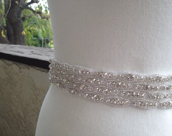 Op de afbeelding: Een close-up van een witte kralenriem met sprankelende strass-steentjes. De riem is gemaakt van meerdere rijen kleine, heldere kralen en grotere, glinsterende strass-steentjes. De riem wordt gedragen rond een wit kledingstuk.