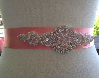May include: A coral pink satin sash with an ornate, beaded embellishment. The central design features a cluster of pearls and clear crystals, surrounded by smaller beads. The sash is tied in a bow.