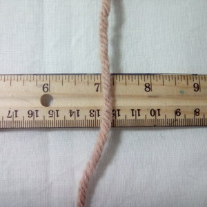 May include: A close-up of a beige yarn strand against a white background. A wooden ruler with black markings is positioned beneath the yarn. The ruler shows numbers 5 through 9, with the yarn strand crossing over the number 7.
