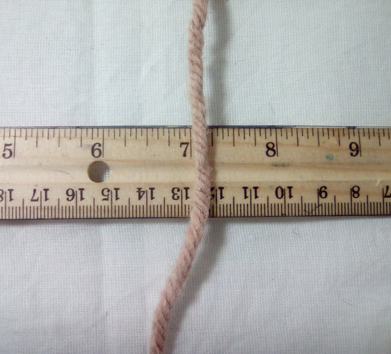 May include: A close-up of a beige yarn strand against a white background. A wooden ruler with black markings is positioned beneath the yarn. The ruler shows numbers 5 through 9, with the yarn strand crossing over the number 7.