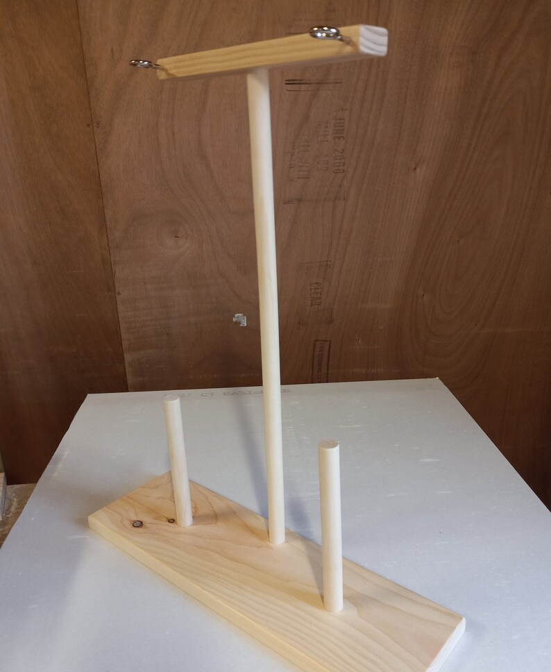 May include: Wooden yarn holder with three dowels and a top bar. The holder is made of natural wood and has a simple design.