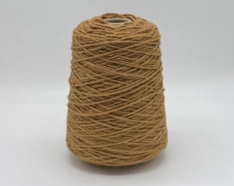 May include: A cone of tan-colored yarn, tightly wound and tapered. The yarn appears to be a natural fiber, suitable for knitting or crocheting projects. The cone is set against a plain white background.