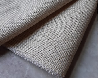 May include: A piece of beige fabric with a visible, coarse, and uniform woven grid pattern. The material is folded over itself, displaying a thick, textured weave with a raw, unfinished edge showing loose threads along the bottom border.