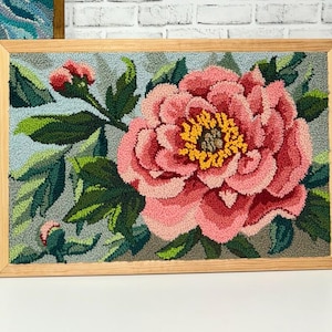 Peut inclure: Une œuvre d'art en point d'aiguille encadrée représentant une grande pivoine rose avec un centre jaune. La fleur est entourée de feuilles vertes et de boutons sur un fond bleu clair.