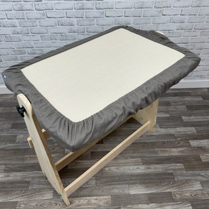 May include: A grey padded changing mat with a white fabric top, attached to a wooden frame with a safety latch. The changing mat is on a wooden stand with a shelf.